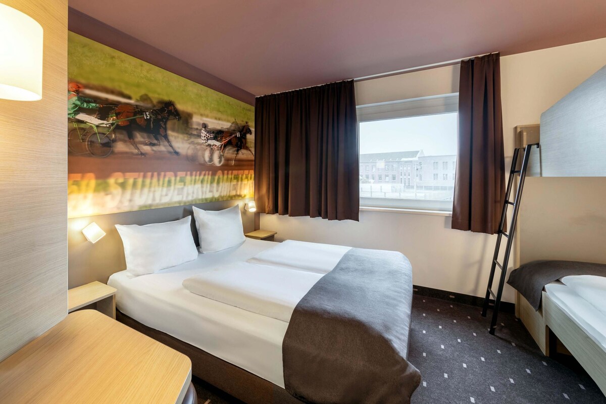 A hotel room with a bed, a desk, a chair, a window with curtains, and a wall mural.