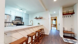 A modern kitchen with white cabinets, a stainless steel refrigerator, a microwave, and a sink. There are wooden stools at the counter and a wooden bench in the adjacent area. - Pet-Friendly. Updated, Private Gas Grill, Located in the Quiet Meadow Area (Mammoth Lakes)