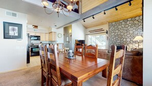 Un grand espace à manger avec une table et des chaises en bois, une cuisine à l’arrière-plan et une cheminée en pierre dans le salon.