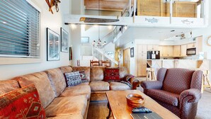 Un vaste espace de vie comprenant un grand canapé d’angle, une table basse en bois et un fauteuil à motifs. Un escalier mène à un niveau supérieur où se trouve un coin cuisine.
