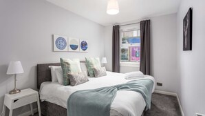 Un dormitorio con una cama, una mesita de noche, una lámpara, una ventana con cortinas y decoración en la pared.