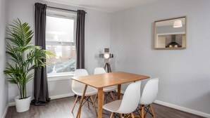Un comedor moderno con una mesa de madera y sillas blancas, una planta en maceta, una ventana con cortinas, una lámpara de pie y un espejo en la pared.