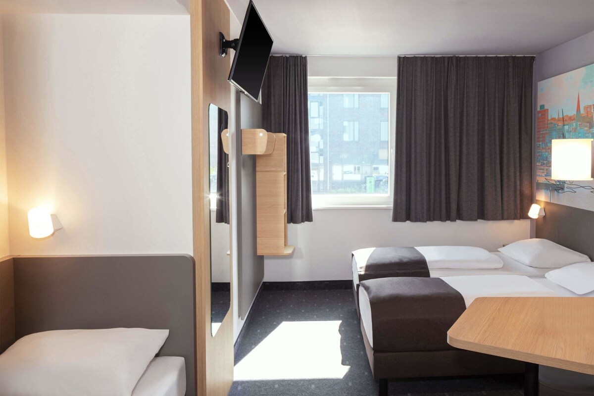 A hotel room with a bed, a desk, a chair, a TV mounted on the wall, and a window with curtains.