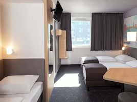 Habitación de hotel con cama, escritorio, silla, televisor montado en la pared y ventana con cortinas.