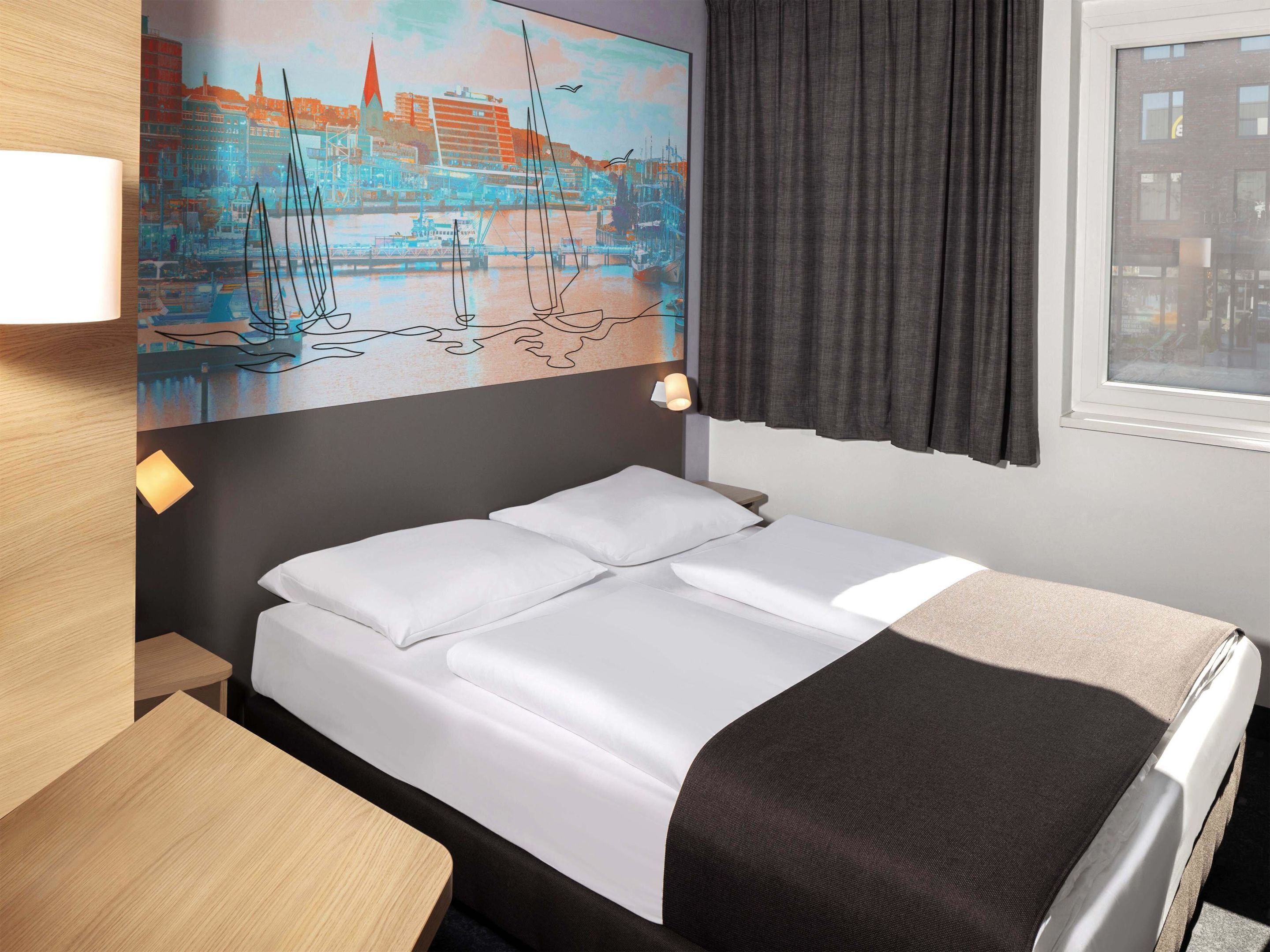 A hotel room with a bed, a desk, a painting of a cityscape, and a window with curtains.