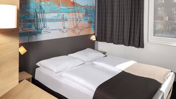 A hotel room with a bed, a desk, a painting of a cityscape, and a window with curtains.