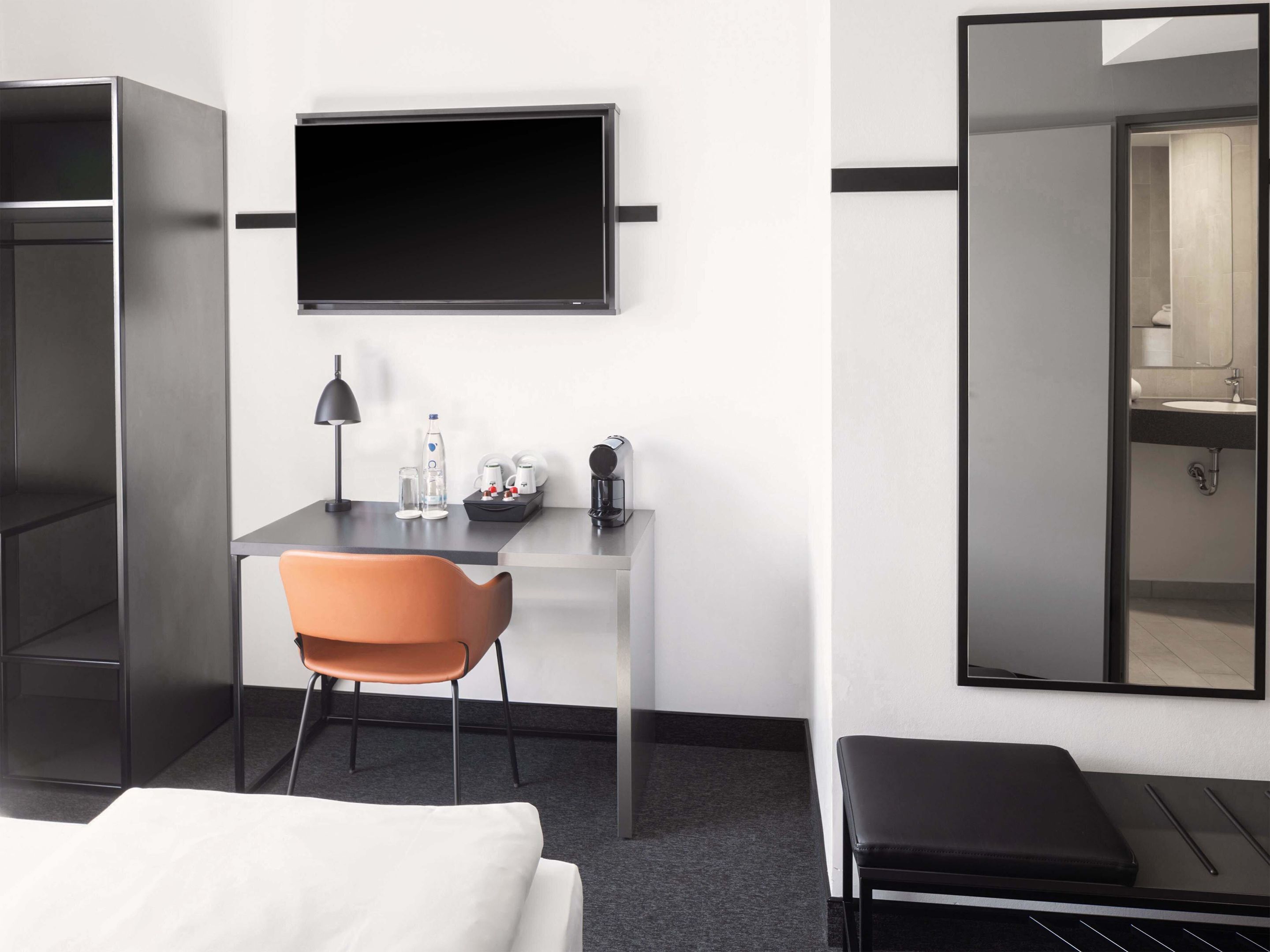 A modern hotel room with a bed, a desk with a chair, a flat-screen TV, a mirror, and a bathroom visible through an open door.