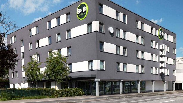 Un moderno hotel tipo B&B con fachada plana, grandes ventanales y un llamativo logo verde y blanco.