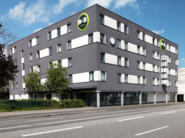 Un moderno hotel tipo B&B con fachada plana, grandes ventanales y un llamativo logo verde y blanco.