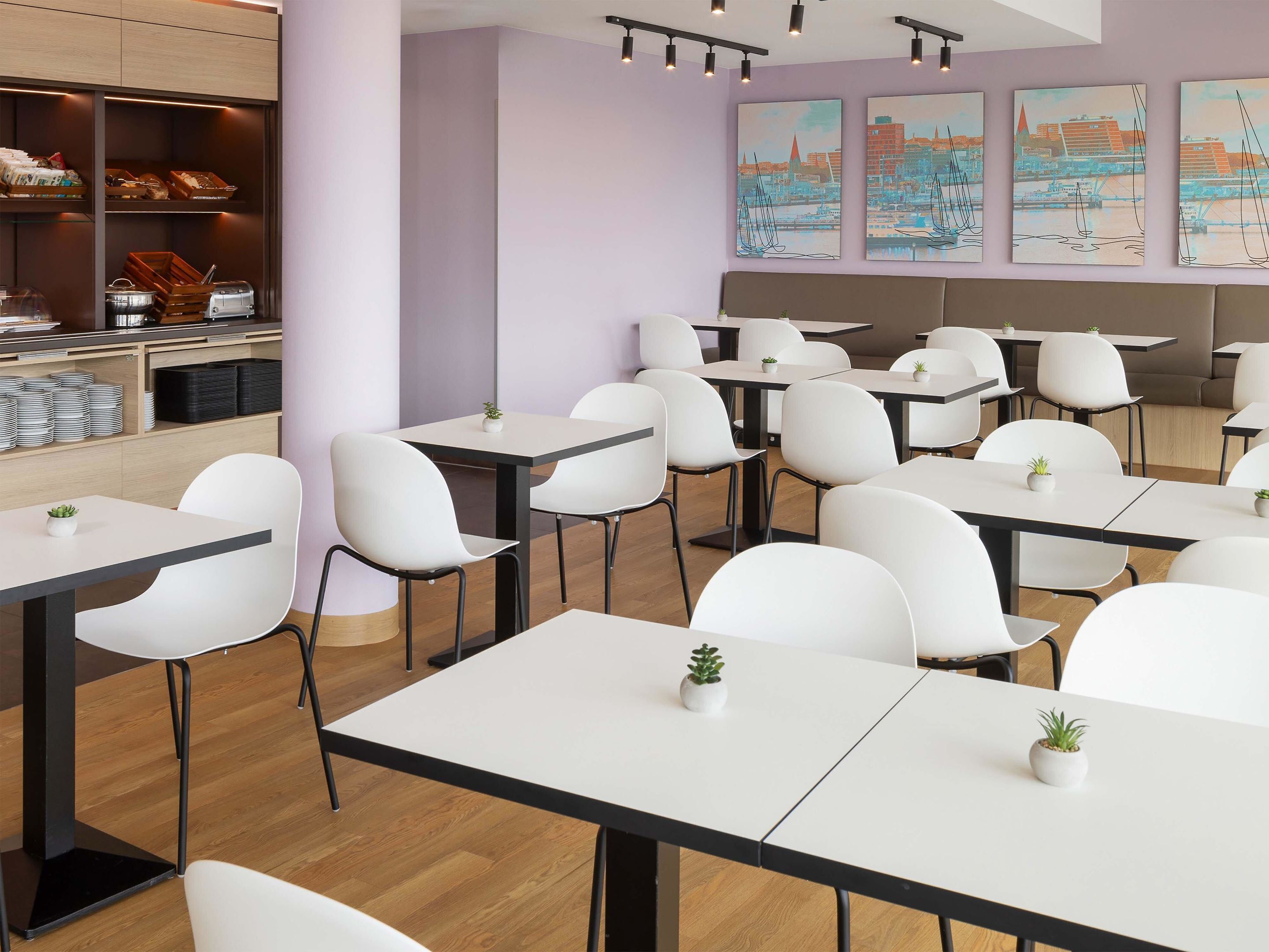 Un espace de restauration moderne avec des chaises et des tables blanches, un comptoir où sont exposées des pâtisseries et des tableaux représentant des paysages urbains accrochés au mur.