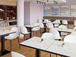 Un comedor moderno con sillas y mesas blancas, una barra con productos de repostería y pinturas de la ciudad en la pared.
