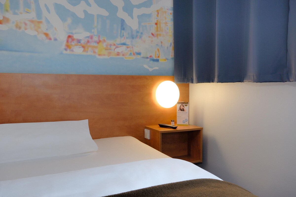 Una habitación de hotel con una cama, una mesita de noche con lámpara, una obra de arte en la pared y un ventanal con cortinas.