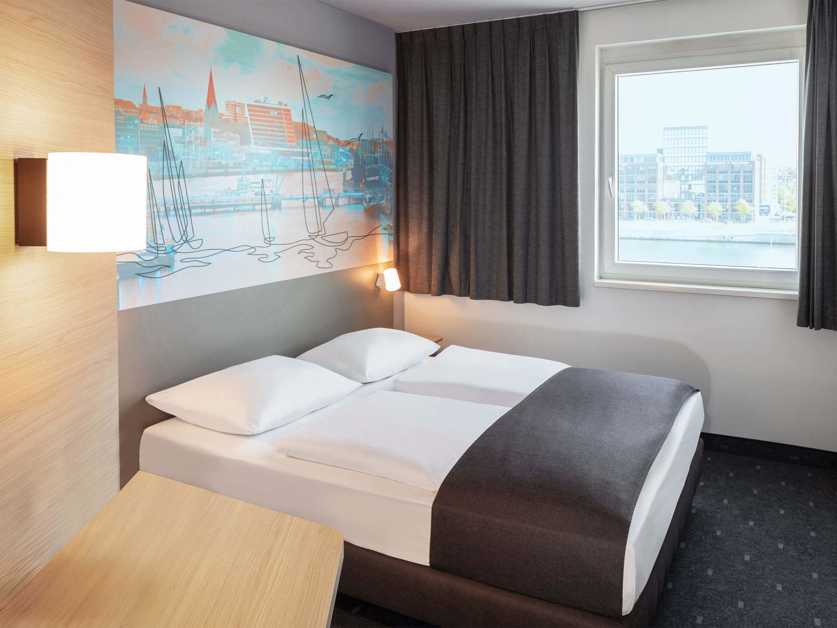 Une chambre d’hôtel avec un lit, un bureau, un tableau représentant un paysage urbain, une fenêtre avec des rideaux et une lampe.
