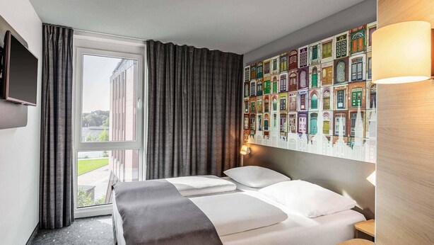 A hotel room with a bed, a TV, a window with curtains, and a wall with colorful building illustrations. - B&B HOTEL Lübeck-Hbf (Lübeck)