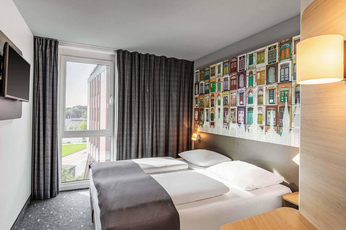 A hotel room with a bed, a TV, a window with curtains, and a wall with colorful building illustrations.