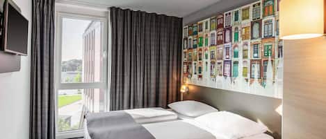 A hotel room with a bed, a TV, a window with curtains, and a wall with colorful building illustrations.