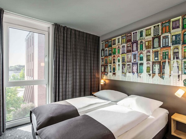 A hotel room with a large window, a bed with white and gray bedding, and a wall mural of colorful building facades. - B&B HOTEL Lübeck-Hbf (Lübeck)