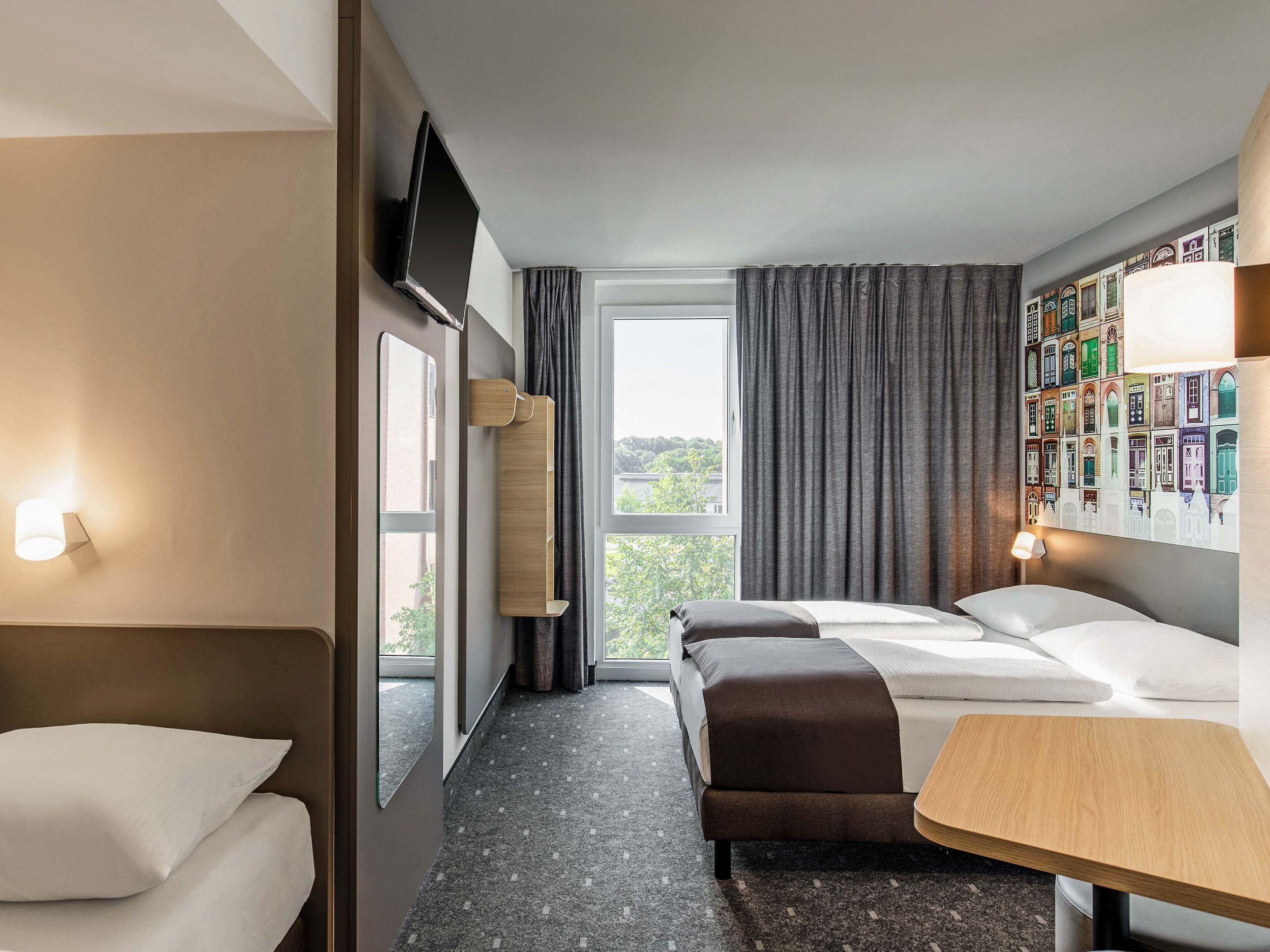 Habitación de hotel con dos camas, una mesita, televisor montado en la pared y una ventana con cortinas.
