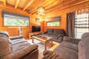Ein gemütliches Blockhaus-Interieur mit Holzbalkendecke, einem Flachbildfernseher, grauen Sofas und einem Couchtisch.