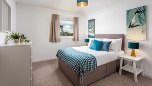 A bedroom with a bed, bedside tables, a window with curtains, a hanging light fixture, and wall art. - Walker Suite No53 - Donnini Apartments (Kilmarnock)