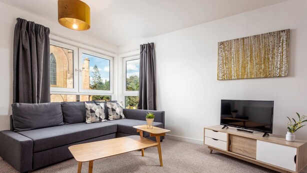 A modern living room with a grey sofa, a wooden coffee table, a TV on a wooden cabinet, and a large window with curtains. - Walker Suite No53 - Donnini Apartments (Kilmarnock)