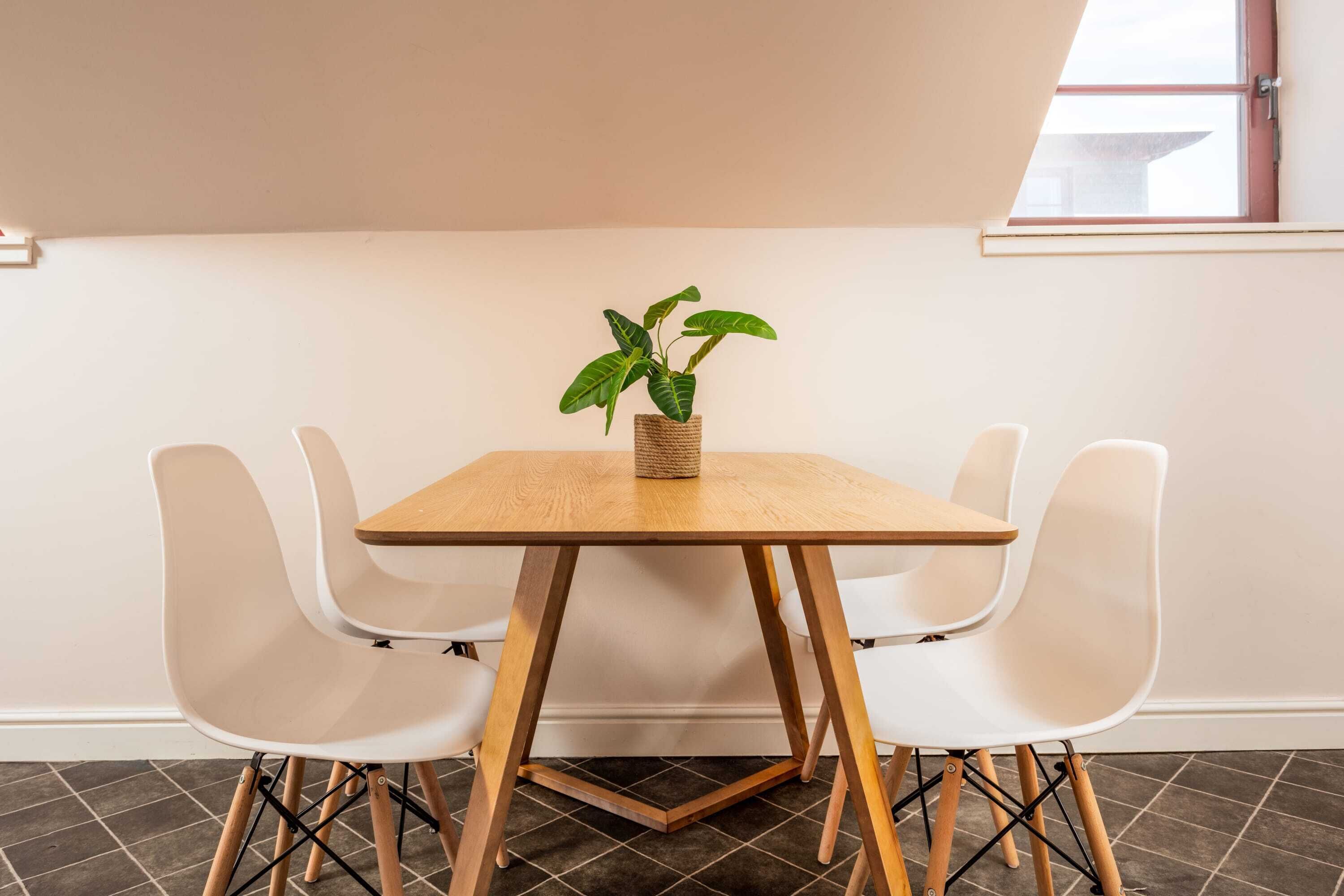 A dining area with a wooden table and white chairs, a potted plant on the table, and a window with a view.