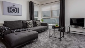A modern living room with a grey sofa, black and white patterned pillows, a small black side table with a plant, a black TV stand, and a large window with curtains. - Walker Suite No73 - Donnini Apartments (Kilmarnock)