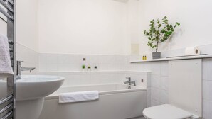 A bathroom with a white sink, bathtub, and toilet. There is a plant on the wall and a shelf with toiletries. - Walker Suite No73 - Donnini Apartments (Kilmarnock)