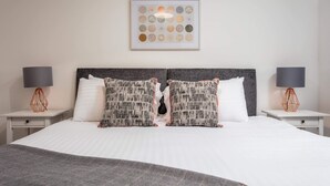 A neatly arranged hotel room with a large bed, two nightstands, a framed artwork above the bed, and a grey headboard. - Walker Suite No73 - Donnini Apartments (Kilmarnock)