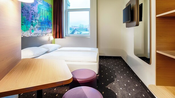 Une chambre d’hôtel équipée d’un lit, d’un bureau, d’une chaise, d’une télévision et d’une fenêtre donnant sur des immeubles.