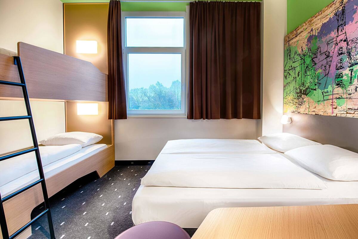 Une chambre d’hôtel avec un lit superposé, un lit simple, une fenêtre avec des rideaux, une carte accrochée au mur et un petit tabouret.