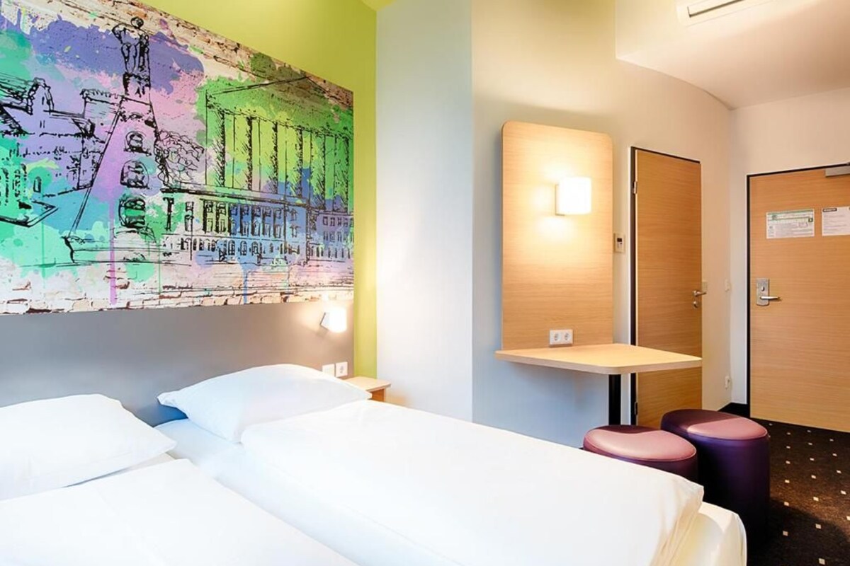 Une chambre d’hôtel avec deux lits, une petite table, une lampe fixée au mur, une porte et une fresque colorée sur le mur.