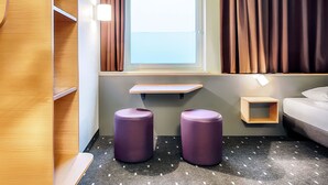A hotel room with a window, curtains, a bed, a small table, and two purple stools. - B&B Hotel Kassel-City (Kassel)