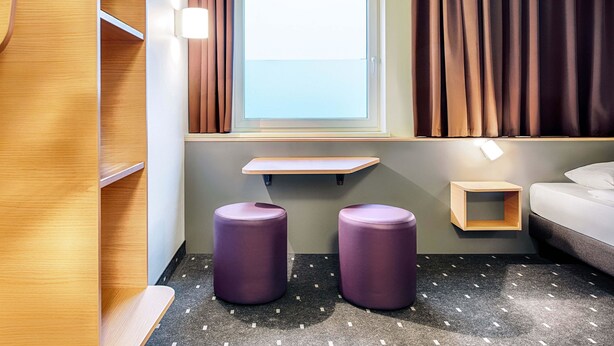 Une chambre d’hôtel avec une fenêtre, des rideaux, un lit, une petite table et deux tabourets violets.