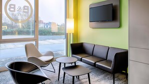 Un hall d’hôtel moderne, doté d’un canapé, de fauteuils, d’un téléviseur et d’une grande fenêtre offrant une vue sur les bâtiments et la verdure.