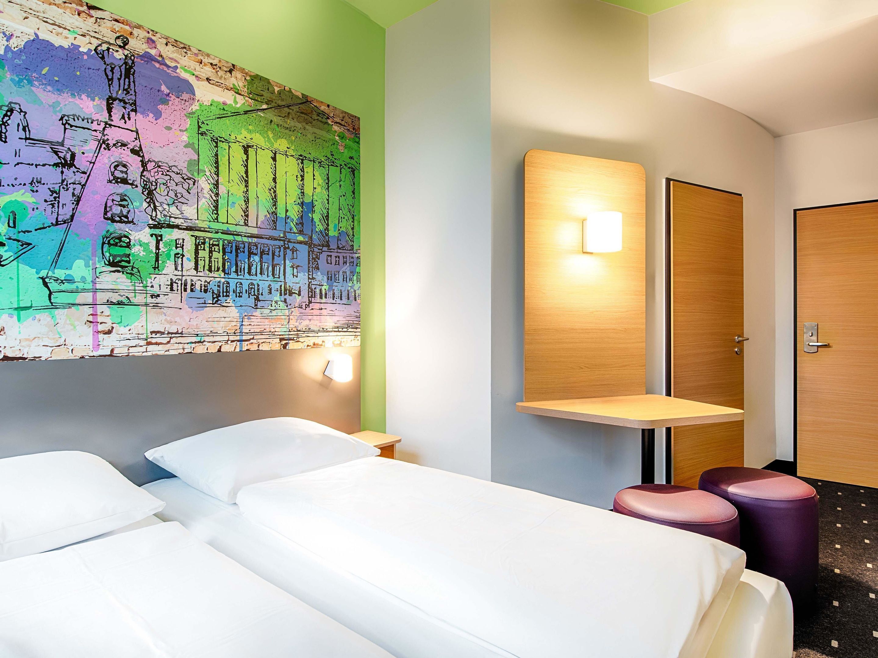 Ein Hotelzimmer mit zwei Betten, einem farbenfrohen Wandgemälde, einem Nachttisch und einer Wandleuchte. Ein Tür ist ebenfalls zu sehen.