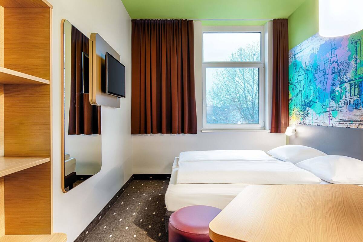 Une chambre d’hôtel avec deux lits, un téléviseur fixé au mur, une fenêtre avec des rideaux, une petite table et une lampe fixée au mur.