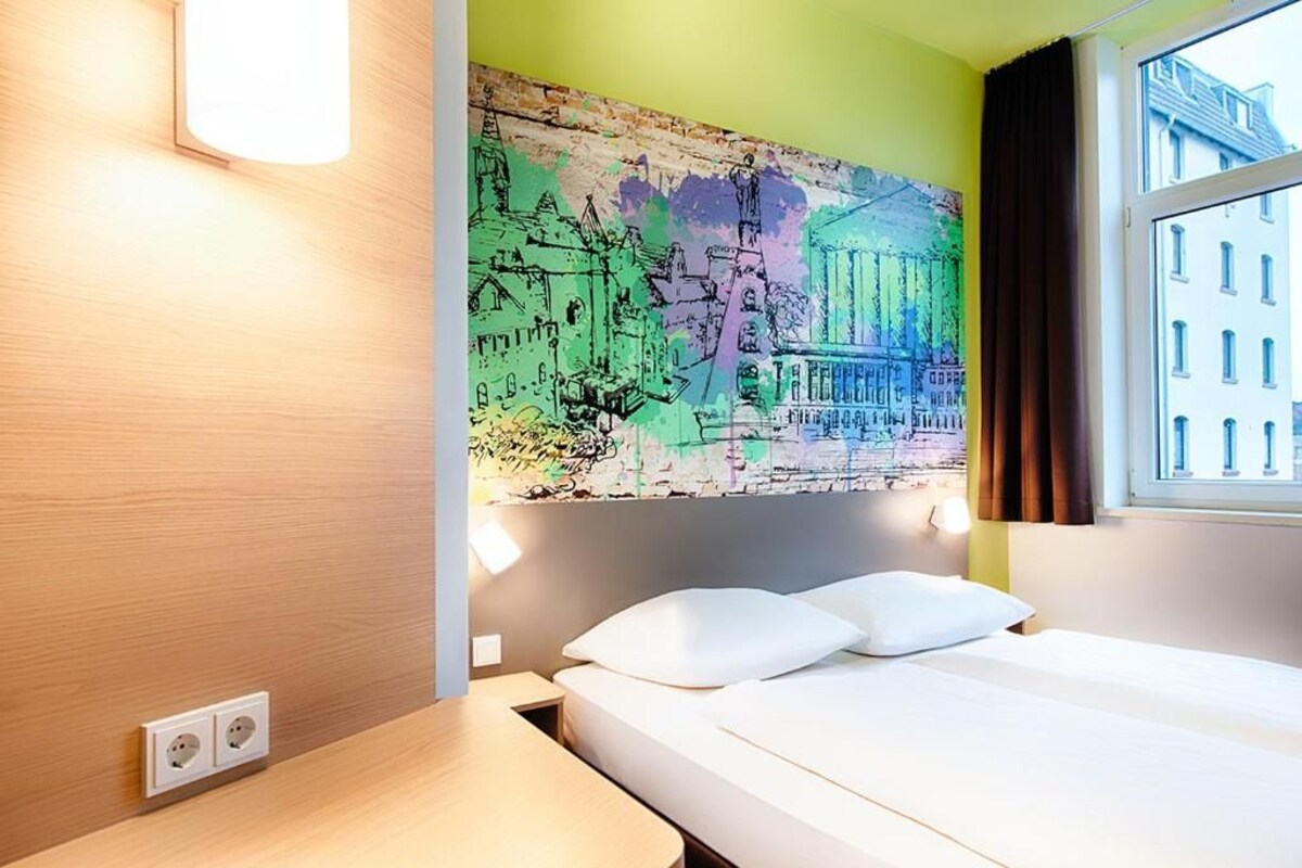 Une chambre d’hôtel avec un lit, une table de chevet, une lampe fixée au mur et une fenêtre avec des rideaux.