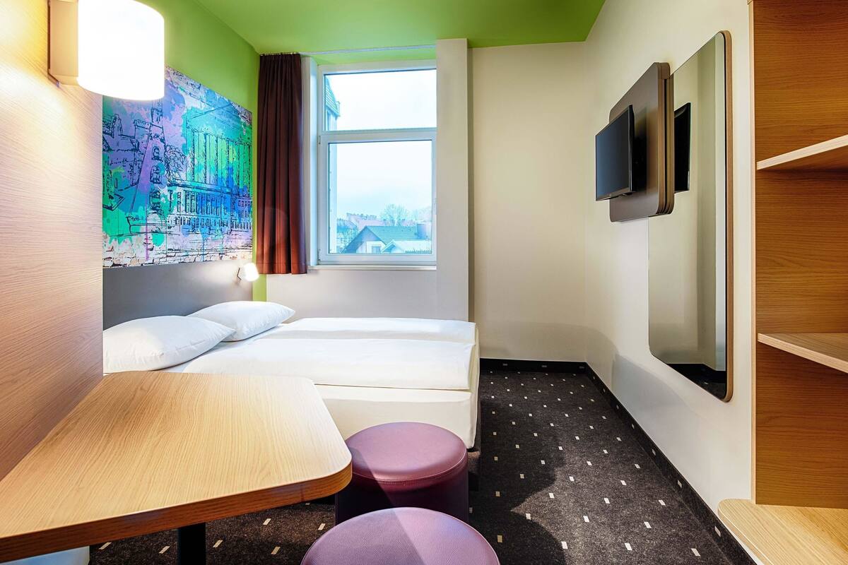 Une chambre d’hôtel équipée d’un lit, d’un bureau, d’une chaise, d’une télévision et d’une fenêtre donnant sur des immeubles.