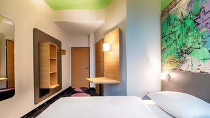 Une chambre d’hôtel avec un lit, un miroir, une étagère et une lampe fixée au mur.