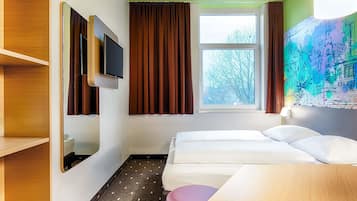 Ein Hotelzimmer mit zwei Betten, einem an der Wand montierten Fernseher, einem Fenster mit Vorhängen, einem kleinen Tisch und einer Wandleuchte.