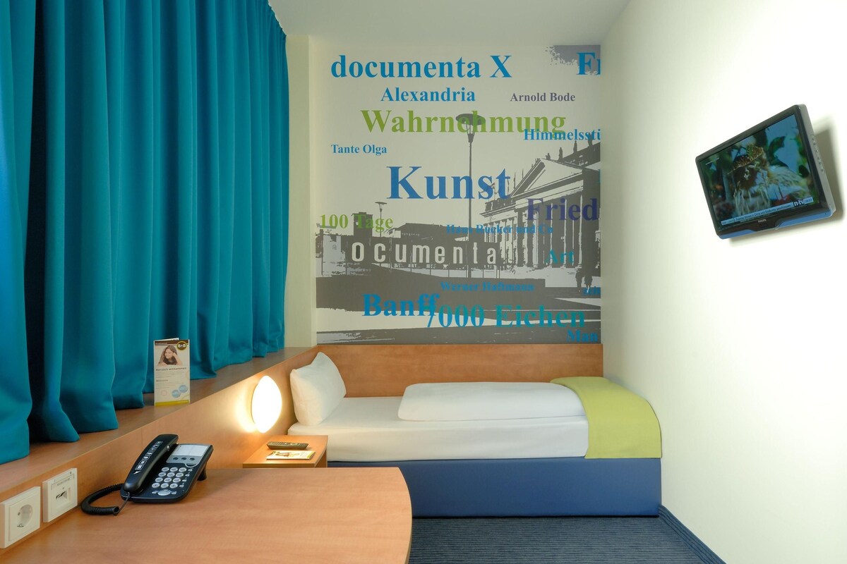 Une chambre d’hôtel avec un lit, un bureau avec un téléphone, un mur orné d’une affiche et un cadre photo.