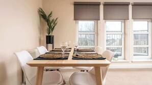 Un coin repas avec une table dressée pour quatre personnes, des chaises blanches, une plante en pot et une fenêtre avec des stores.