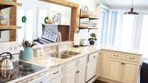 Une cuisine avec des meubles blancs, un plan de travail en granit et une cuisinière. Il y a des étagères au-dessus de l’évier et une plante en pot sur le plan de travail.
