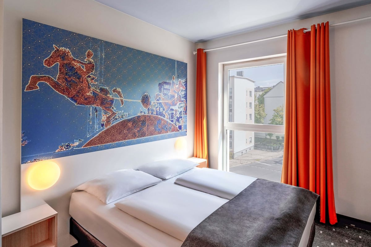 Ein Hotelzimmer mit Bett, einem Wandbild, einem Fenster mit Vorhängen und Blick ins Freie.