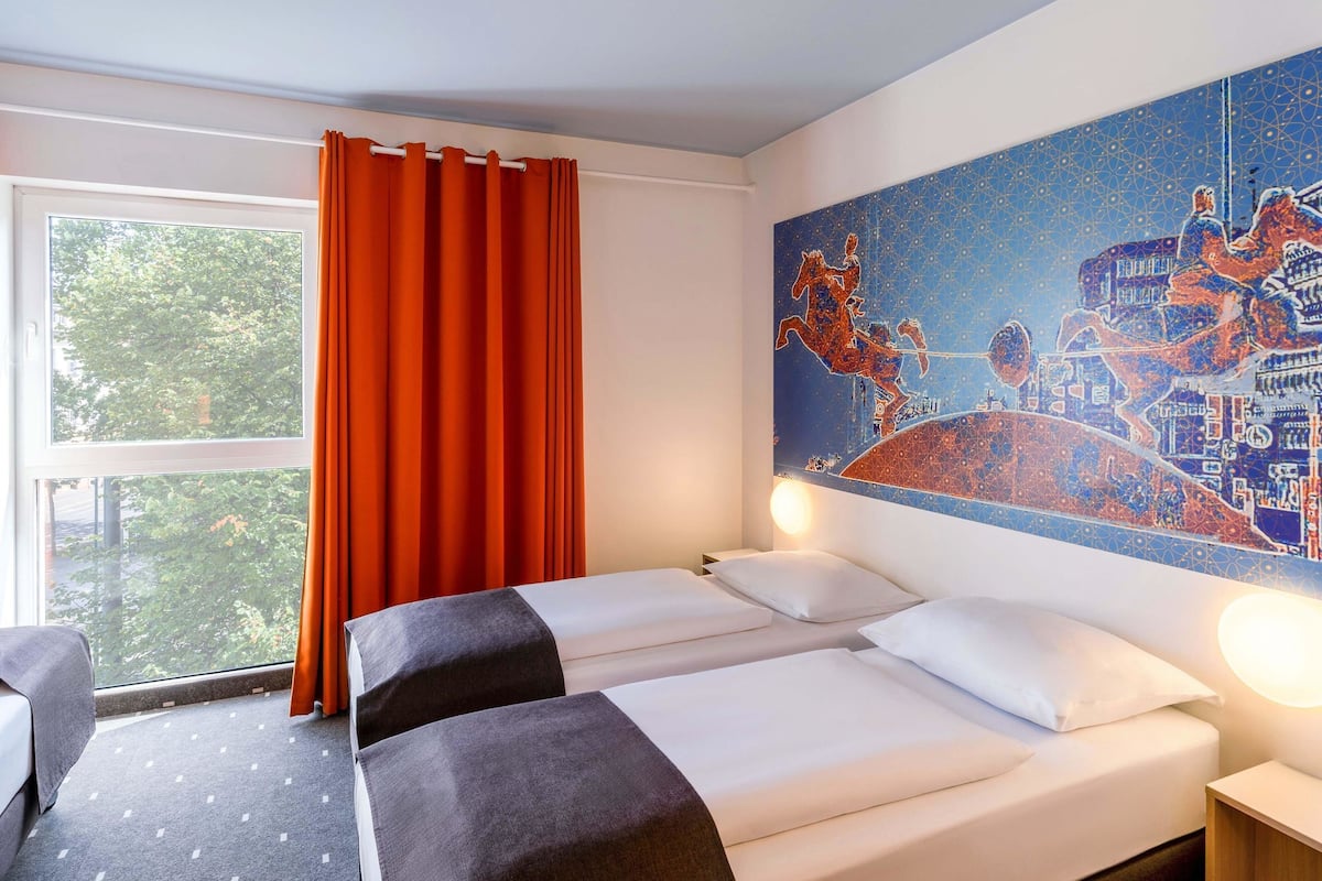 Ein Hotelzimmer mit zwei Betten, einem großen Fenster mit orangefarbenen Vorhängen und einem Wandgemälde, das einen Reiter und sein Pferd zeigt.