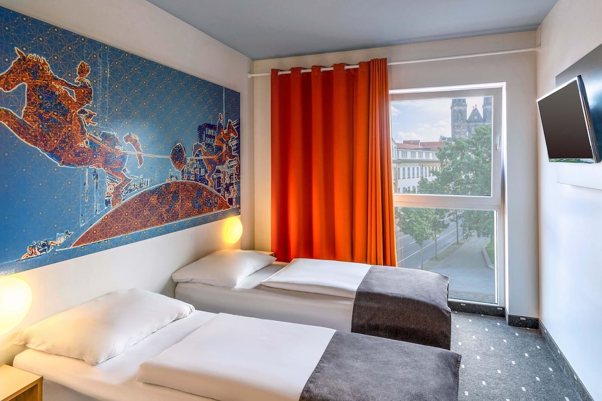 Ein Hotelzimmer mit zwei Betten, einem großen Fenster mit orangefarbenen Vorhängen, einem Stadtbild-Wandgemälde und einem an der Wand befestigten Flachbildfernseher.