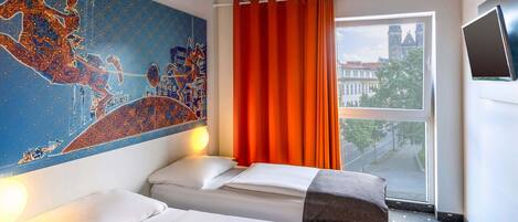 Habitación de hotel con dos camas, un ventanal con cortinas naranjas, un mural de paisaje urbano en la pared y un televisor de pantalla plana montado en la pared.