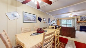 Un comedor con mesa de madera, sillas y un cuenco de piñas. Hay una chimenea y una ventana con cortinas.
