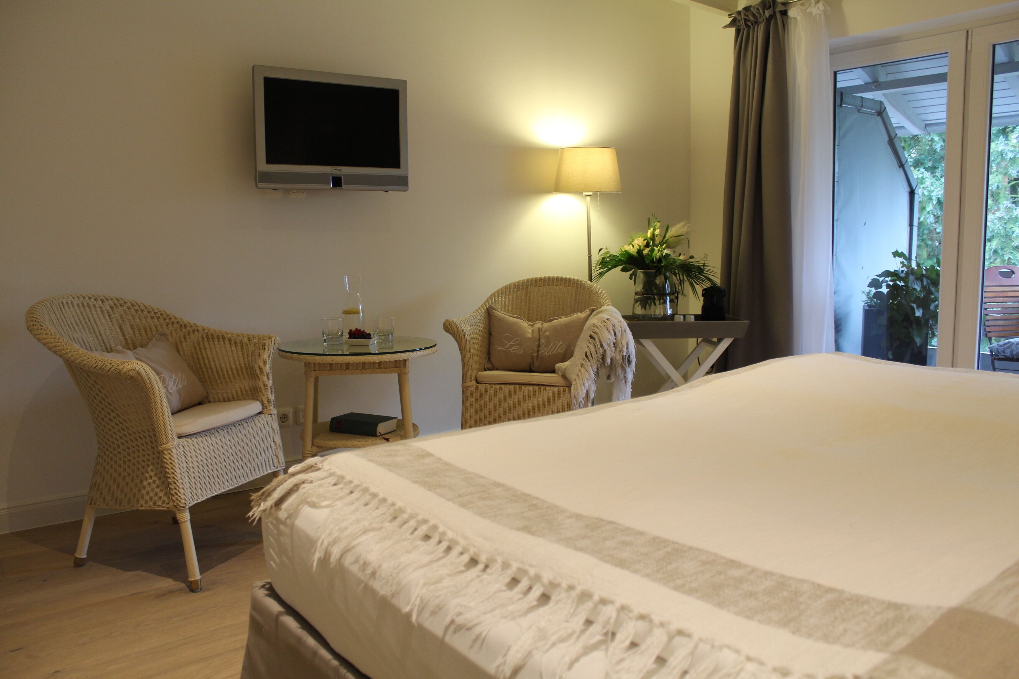 A hotel room with a large bed, two wicker chairs, a small table with a lamp, and a TV on the wall.